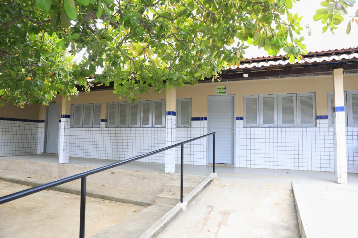 Fachada de uma escola com paredes de azulejos brancos e azuis, janelas cinzas e árvores verdes.